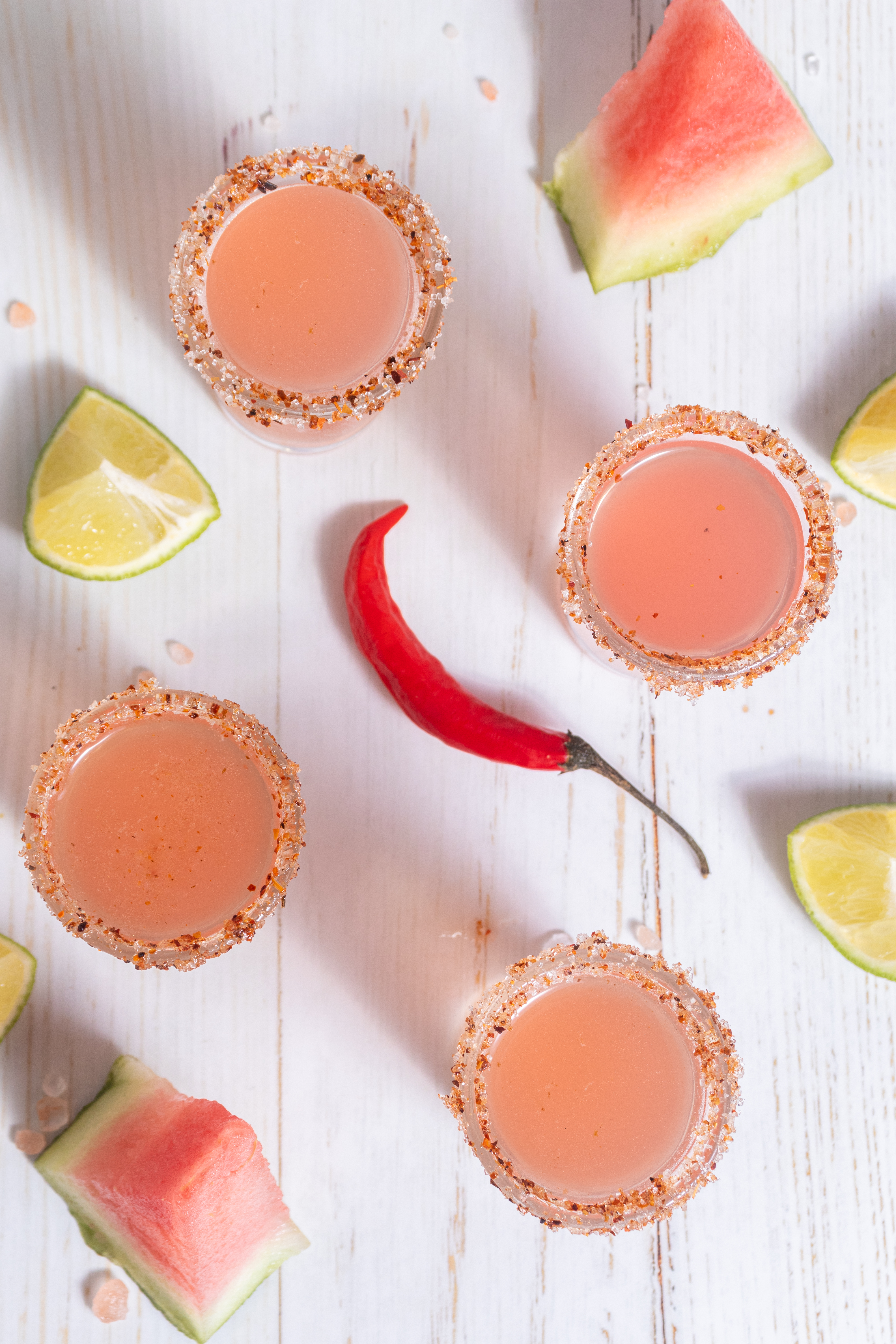 Mexican candy shots rimmed with tajin and sugar next to lime and watermelon wedges and a chili pepper